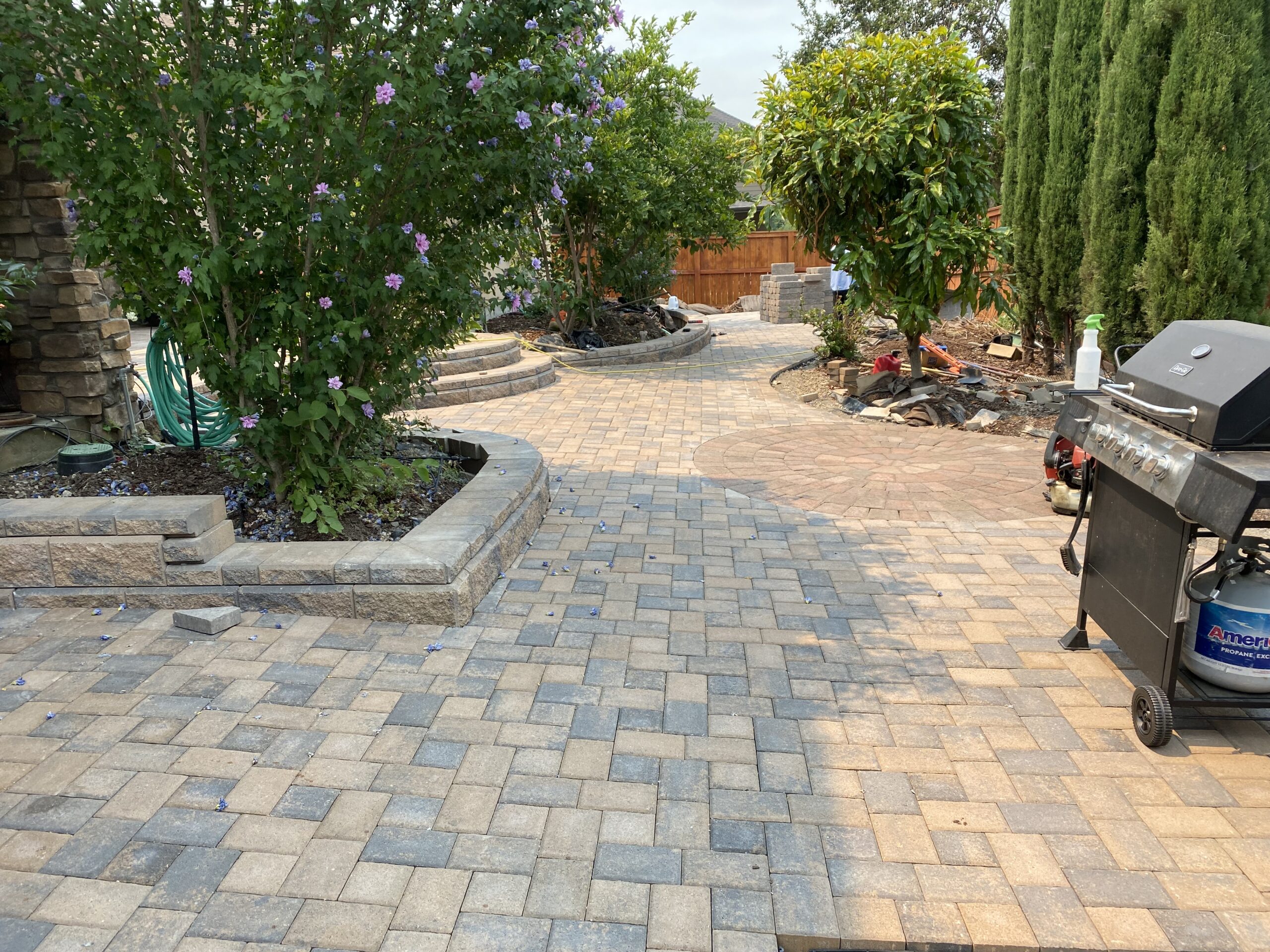 Elegant backyard patio with interlocking stone pavers in hues of tan and gray, bordered by raised garden beds, blooming shrubs, and tall greenery, with a barbecue grill to one side and gardening work in progress in the background.